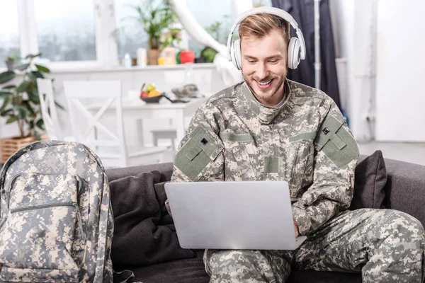 微笑的陆军士兵戴着耳机 在沙发上使用笔记本电脑 — 图库照片