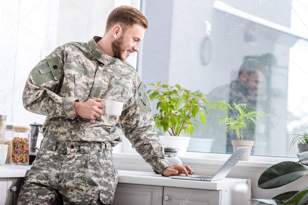 handsome army soldier holding cup of hot coffee and using laptop at kitchen