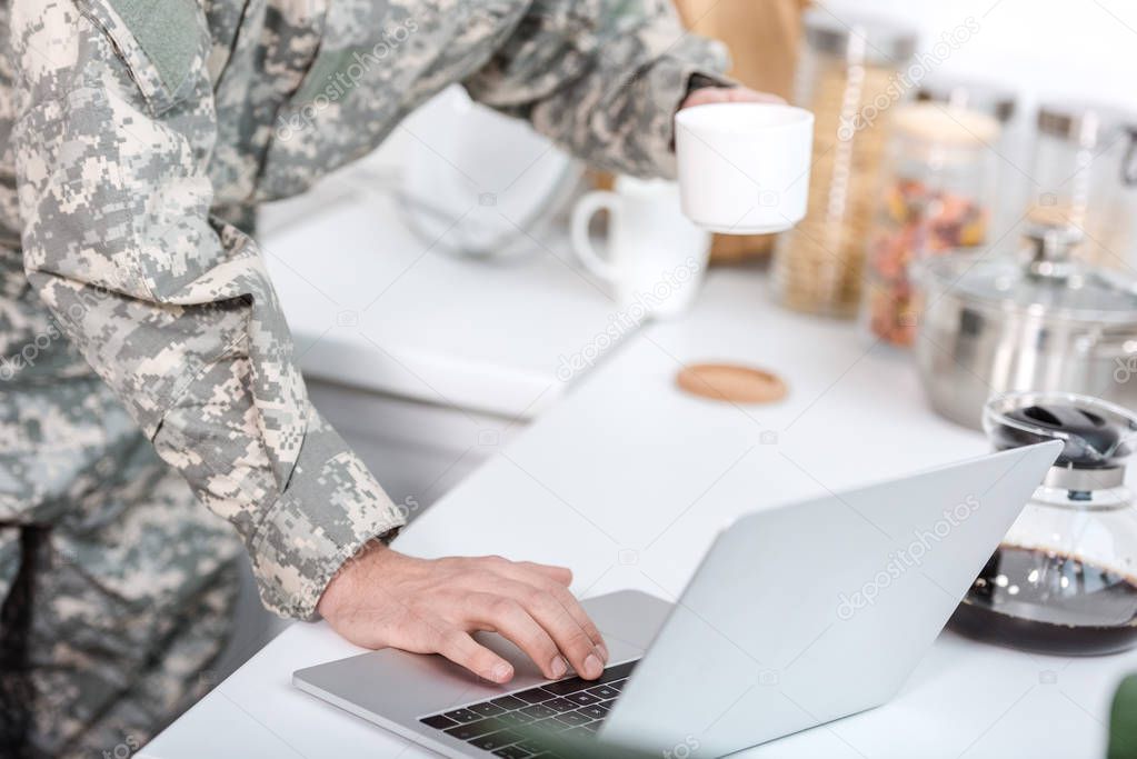 cropped view of soldier holding cup of coffee and using laptop at kitchen