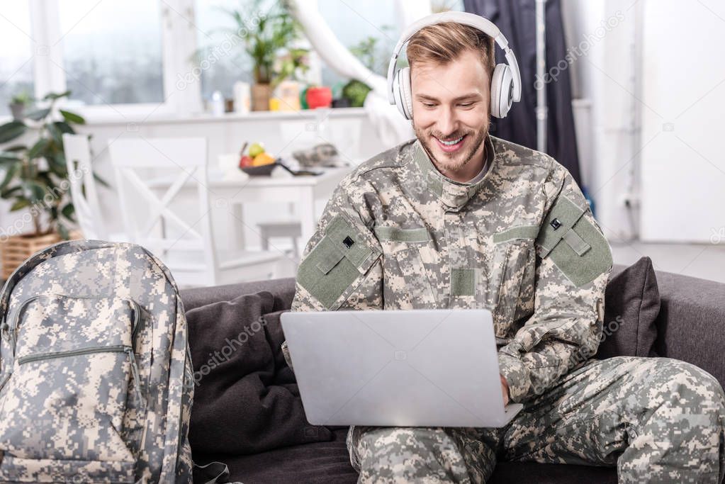 smiling army soldier wearing headphones and using laptop on couch