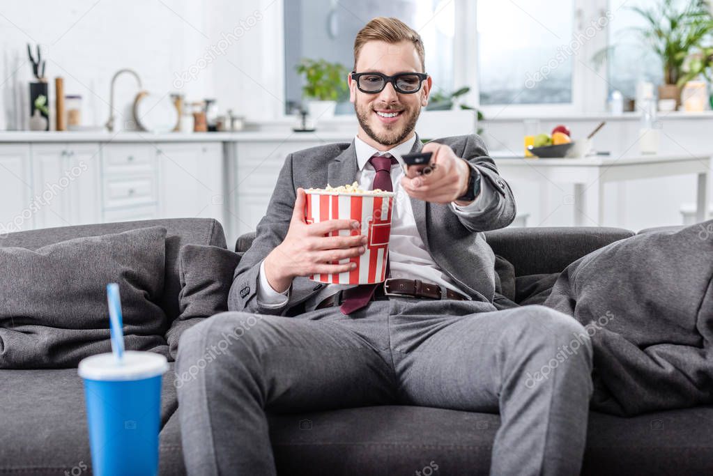 handsome businessman on couch in 3d glasses with remote control watching movie and eating popcorn