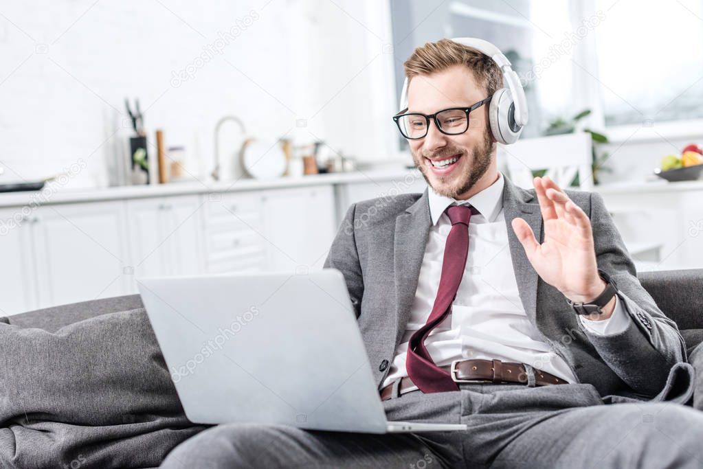 businessman in glasses wearing headphones, waving with hand and using laptop on couch