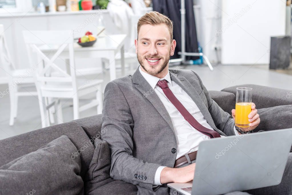 smiling businessman in formal wear drinking orange juice and using laptop