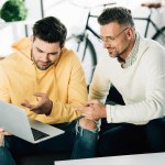 Son showing something on laptop to mature father on sofa at home