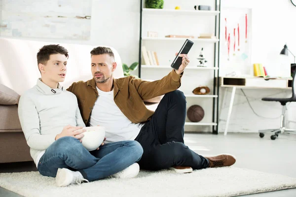 认真的父亲和十几岁的儿子看电视 拿着一碗爆米花在家里 — 免费的图库照片