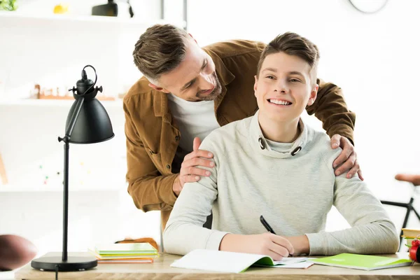 Sonriente Padre Abrazando Feliz Adolescente Hijo Mientras Haciendo Tarea —  Fotos de Stock