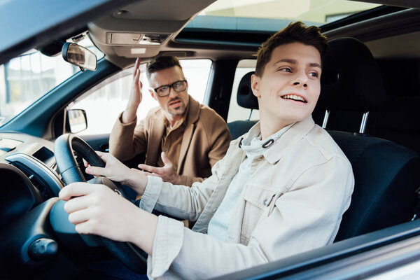 irritated father teaching smiling teen son driving car