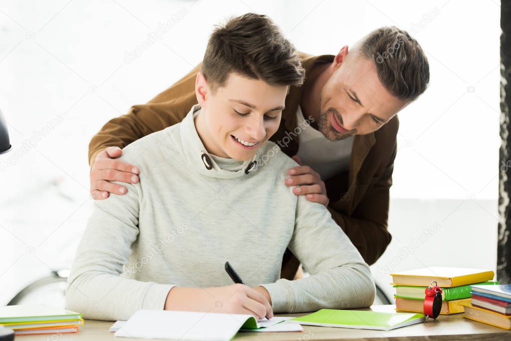 smiling father hugging teen son while he doing homework and writing something