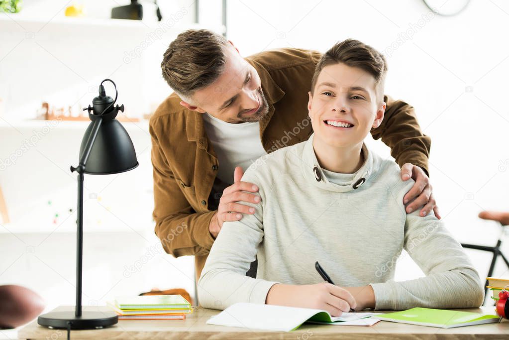 smiling father hugging happy teen son while he doing homework