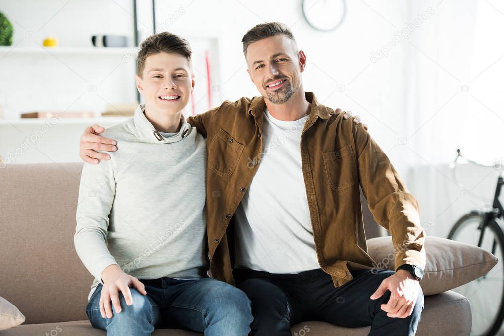 happy father and teen son sitting on sofa and looking at camera at home