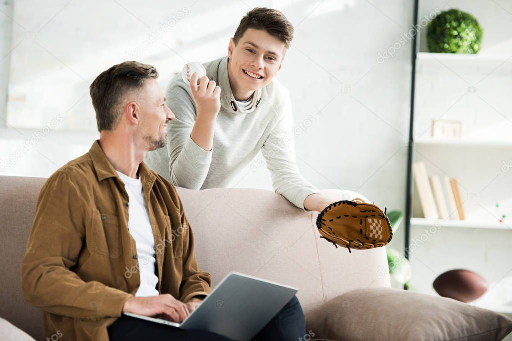 smiling father using laptop and teen son holding baseball glove and ball