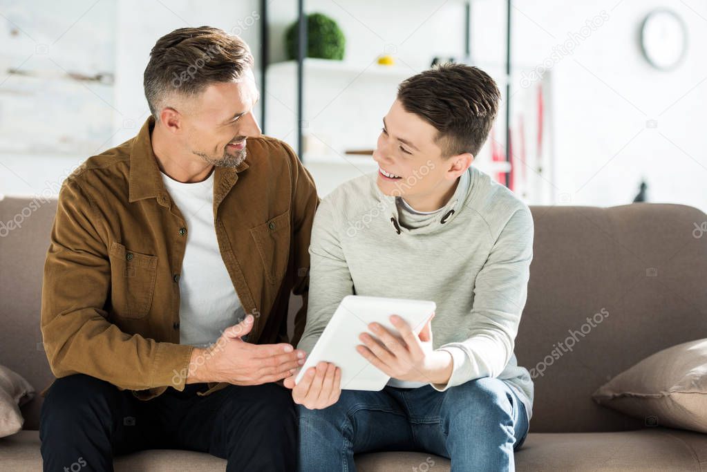 happy father and teen son sitting on sofa with tablet and looking at each other at home