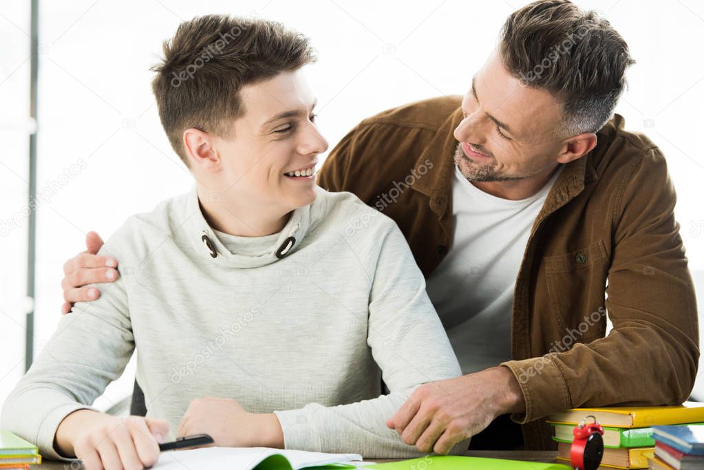 smiling father hugging teen son while he doing homework, looking at each other