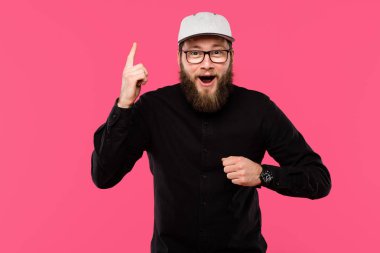 excited bearded man in eyeglasses and cap hat doing idea gesture isolated on pink clipart