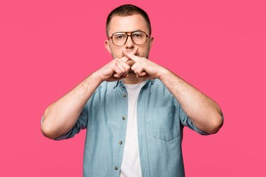 young man in eyeglasses gesturing for silence and holding crossed fingers near lips isolated on pink 