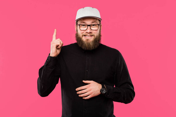 happy bearded man in eyeglasses and cap hat doing idea gesture isolated on pink