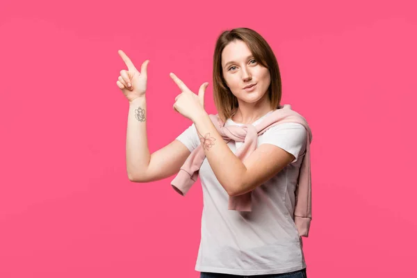 happy tattooed woman pointing by fingers isolated on pink