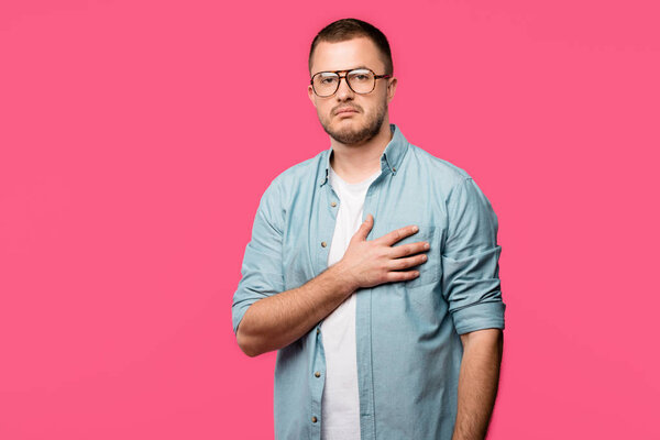 young man in eyeglasses holding hand over heart and looking at camera isolated on pink
