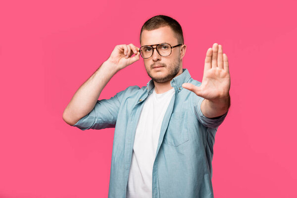 serious man adjusting eyeglasses and showing no sign isolated on pink