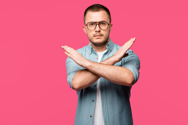 young man gesturing no with crossed arms and looking at camera isolated on pink 