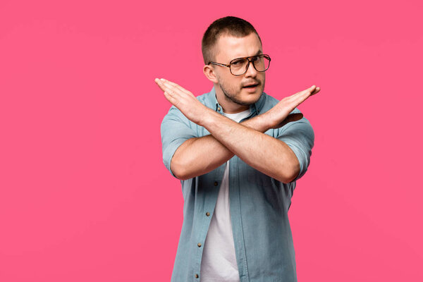 young man in eyeglasses gesturing no with crossed arms and looking at camera isolated on pink 