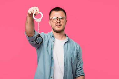 young man in eyeglasses holding handcuffs and looking at camera isolated on pink clipart