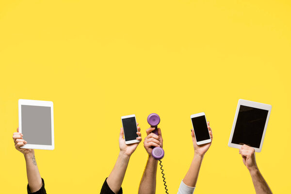 cropped shot of hands holding smartphones, digital tablets and handset isolated on yellow 