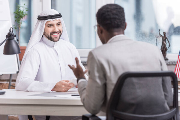 Arabian businessman sitting at desk and looking at african american partner in office 