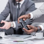 Cropped view of multiethnic businessmen sitting at table with documents and holding gamepads