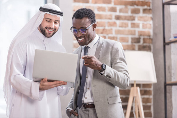 African american businessman poiniting at laptop and smiling near arabian partner 
