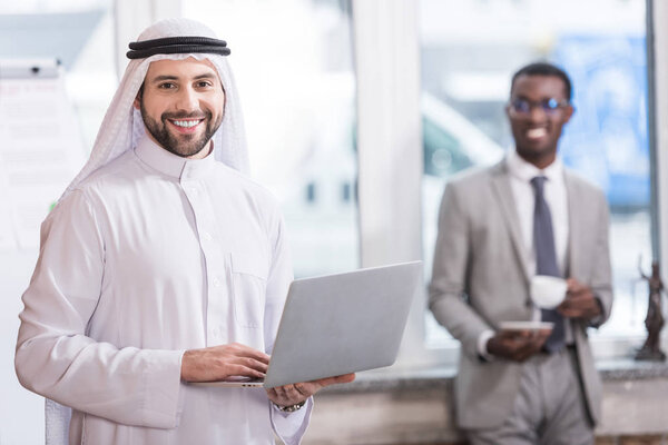 Arabian businessman holding laptop in office with african american partner on background 