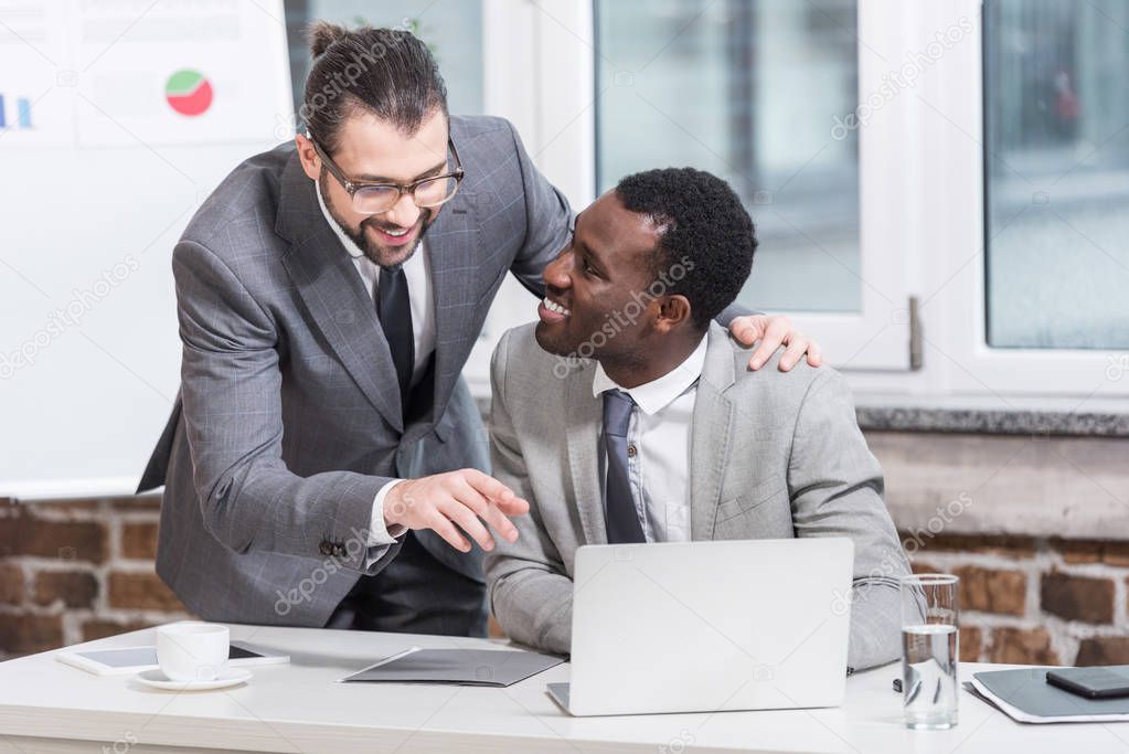 handsome businessman embracing african american partner and pointing at laptop in office