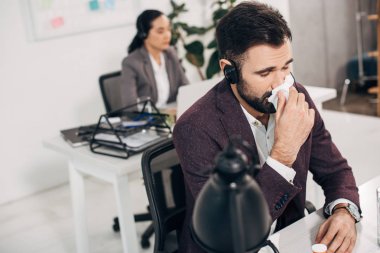 sick call center operator with napkin blowing nose with coworker on background clipart