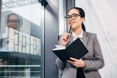 gülümseyen, cam kenarında duran ve defter ve kalem tutan güzel iş kadını
