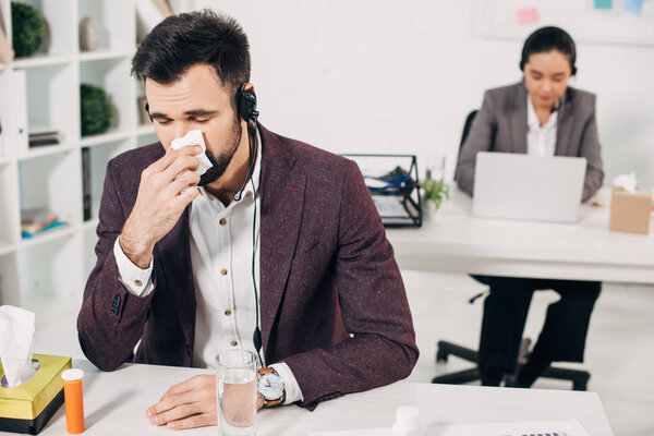 sick call center operator with napkin blowing nose with coworker on background