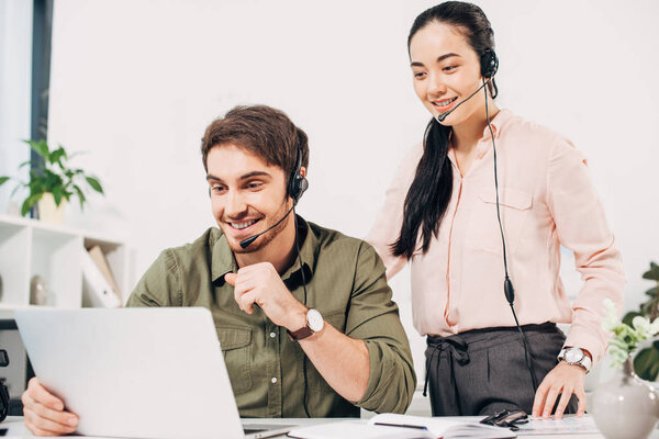 handsome call center operator working at laptop and pretty coworker standing behind in office