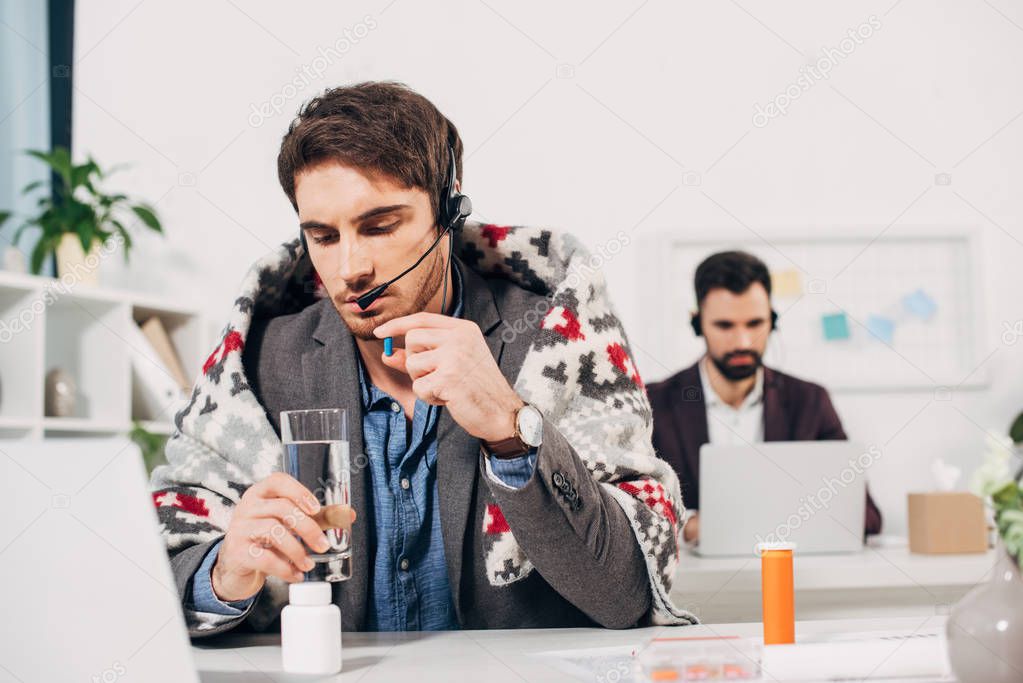 sick call center operator with blanket on shoulders holding glass of water and pill in office