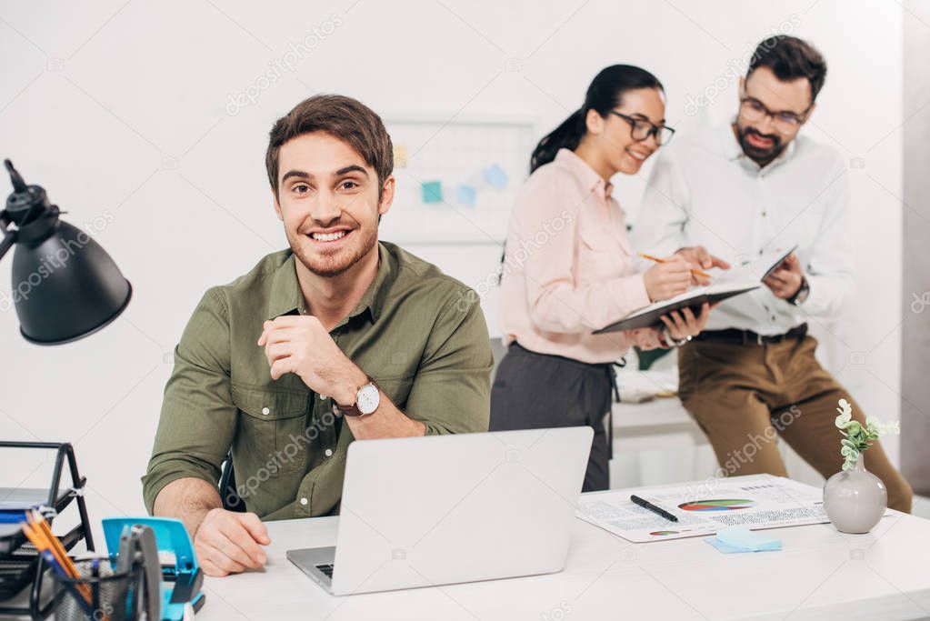 Selective focus of male office manager sitting and smiling with colleagues on background 
