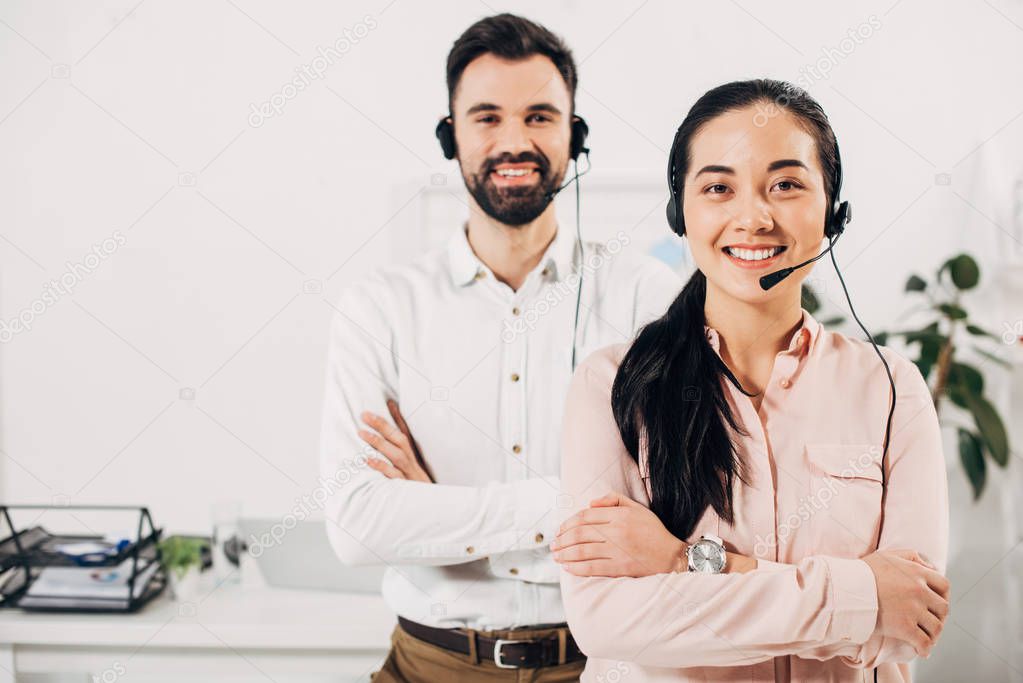 Selective focus of female manager smiling with crossed arms while male coworker standing behind with headset