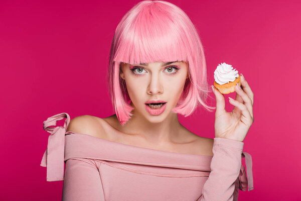 шокированная девушка в розовом парике держа сладкий кекс изолирован на розовый

