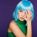 Sensual young woman posing in blue wig and green turtleneck, isolated on purple