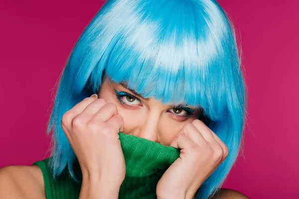 stylish young woman with blue hair hiding face in green turtleneck, isolated on pink