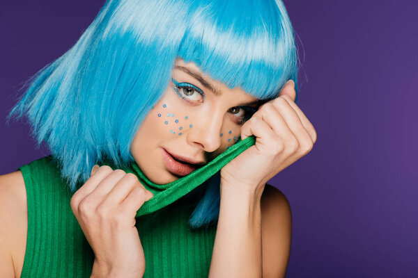 Очаровательная девушка с голубым париком и звездами на лице позируют с зеленой водолазкой, изолированные на фиолетовый
