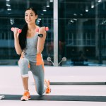 Улыбающаяся азиатская спортсменка делает упражнения с гантелями на фитнес-коврике в спортзале
