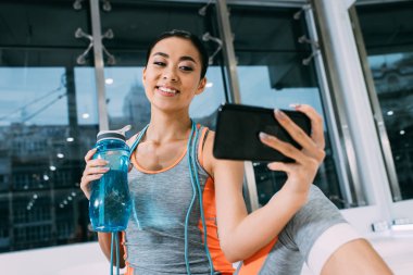 spor şişe su ile tutarak ve selfie spor salonunda alarak gülümseyen Asyalı kız görünümünü kapat