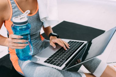 Kırpılan fitness mindere oturmuş, tipik dizüstü klavye üzerinde ve spor salonunda spor şişe tutan kız görünümünü