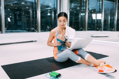 fitness mat dizüstü bilgisayar ile oturma ve spor salonunda spor şişe tutan Asya sporcumuz