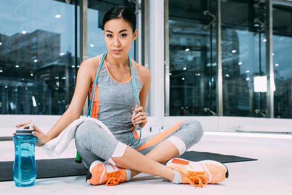 attractive asian girl with skipping rope on shoulders sitting on fitness mat, touching sports bottle with water and looking at camera at gym