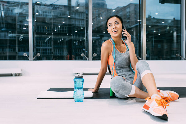 attractive asian sportswoman sitting on fitness mat and talking on smartphone in sports center
