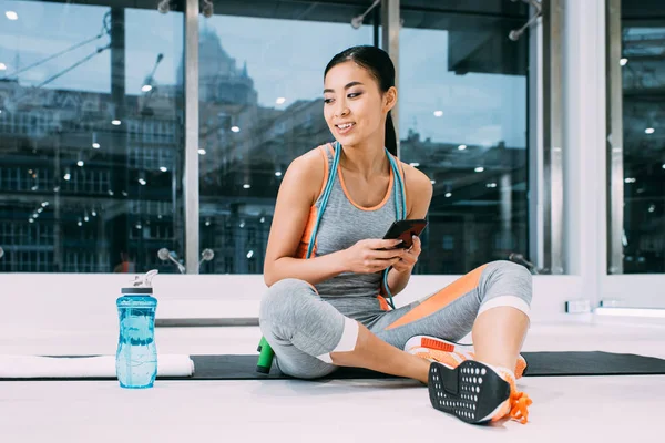 Sorrindo Asiático Esportista Sentado Tapete Fitness Segurando Smartphone Centro Esportes — Fotografia de Stock